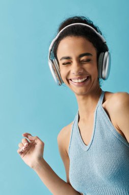 Mavi bir yular giyen genç bir kadın mavi arka planda kulaklıkla müzik dinlerken dans ediyor ve gülümsüyor..