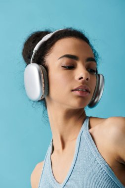 Mavi kolsuz tişörtlü genç bir kadın parlak mavi bir arka planda gümüş kulaklıkla müzik dinliyor..