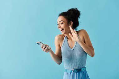 Mavi elbiseli genç bir kadın telefonunu parlak mavi bir arka plana karşı kullanırken geniş bir gülümseme sergiliyor..