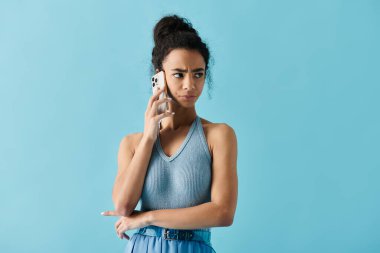 Mavi elbiseli genç bir Afro-Amerikalı kadın açık mavi bir arka planda duruyor ve telefonuyla konuşuyor..