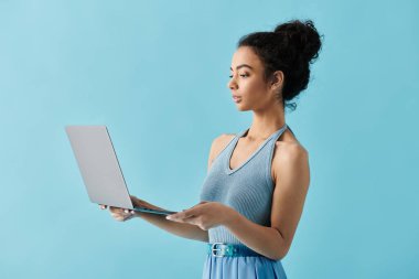 A young woman in a blue dress holds a laptop in front of a light blue background. clipart