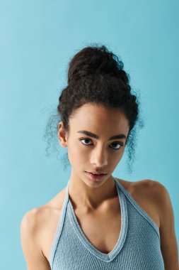 A young African American woman with a confident gaze, wearing a blue halter top, stands against a vibrant blue background. clipart