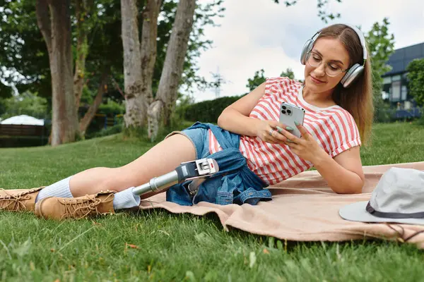 stock image A young woman with a prosthetic leg relaxes in a park, using her phone and listening to music.