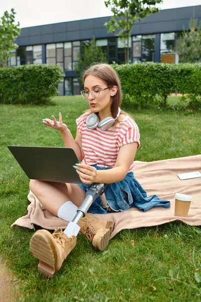 stock image A young woman with a prosthetic leg sits on a blanket in a park, using a laptop and gesturing with her hands.