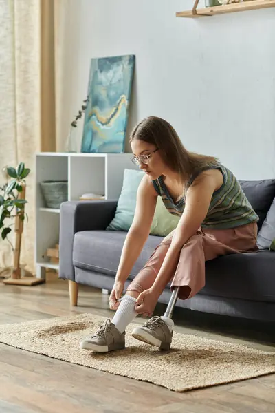 stock image A young woman with a prosthetic leg puts on her shoes while sitting on a sofa in her living room.