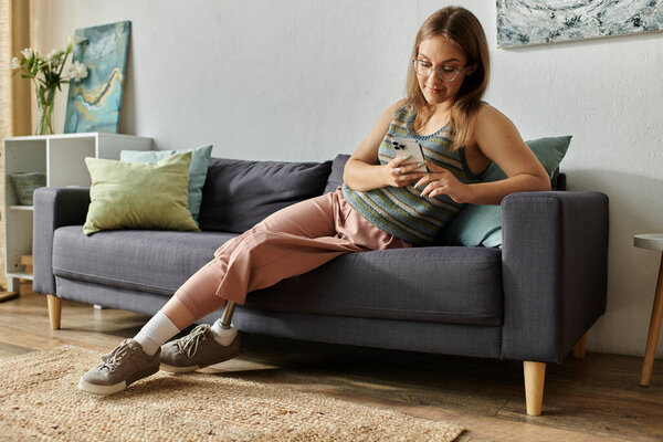 Молодая женщина с протезной ногой сидит на диване в своей гостиной, удобно прокручивая свой телефон.