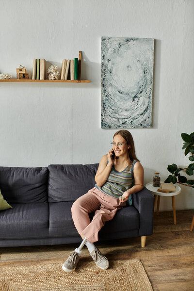 A young woman with a prosthetic leg sits on a couch in her modern living room, smiling as she chats on the phone.