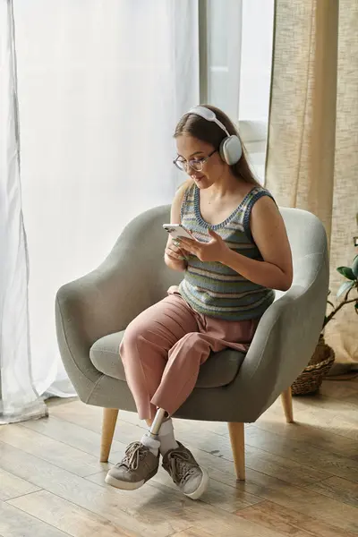 stock image A young woman with a prosthetic leg sits in a comfortable chair, listening to music and scrolling through her phone.