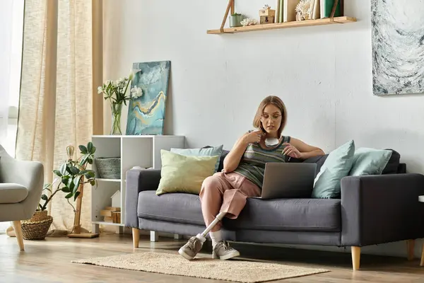 stock image A young woman with a prosthetic leg sits on a couch in her modern living room, using a laptop.