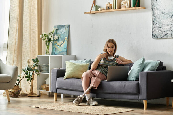 A young woman with a prosthetic leg sits on a couch in her modern living room, using a laptop.