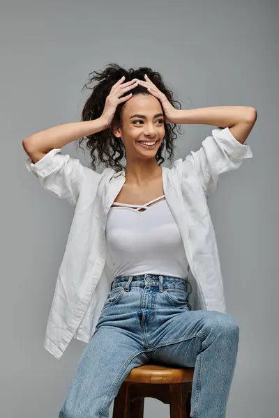 stock image Curly-haired woman in white shirt, jeans, smiles against grey backdrop, looking right while seated on stool