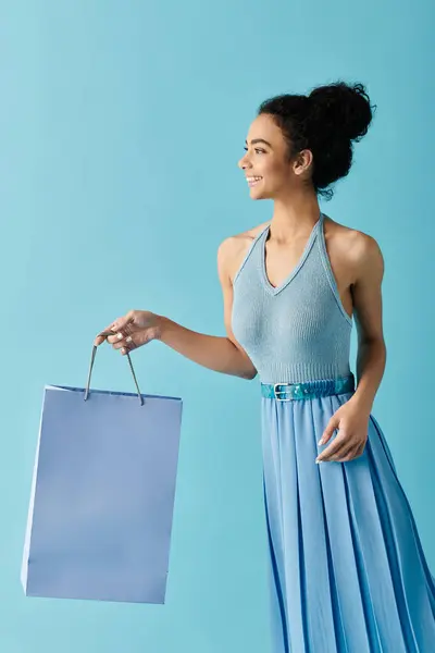 Stock image A young woman in a dress walks with a shopping bag against a bright blue background.