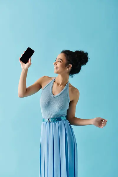 stock image A young woman in a blue dress smiles while holding her phone in front of a light blue background.