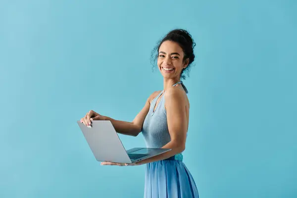 stock image A young woman in a blue dress smiles confidently while holding a laptop in front of a blue background.