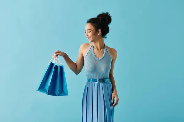 stock image A young woman in a blue dress smiles happily as she holds shopping bags against a blue background.