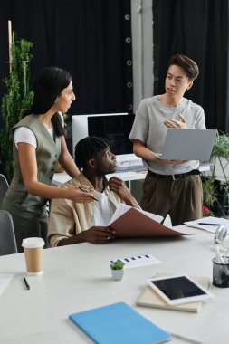 Three colleagues collaborate on a project, reviewing documents and using a laptop. clipart