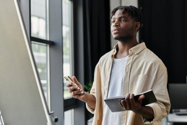 Gündelik giyinmiş genç bir adam, elinde telefon ve defterle modern bir ofis alanında kafa yoruyor..