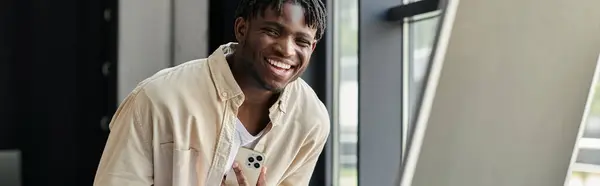 stock image A young man smiles brightly while holding a smartphone in a modern office setting.