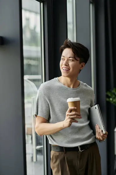 stock image A young man holds a coffee cup and looks out a window in an office building.