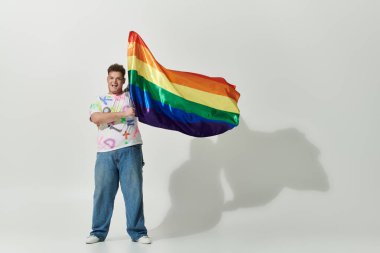 A joyful queer person waves a rainbow flag against a white backdrop. clipart