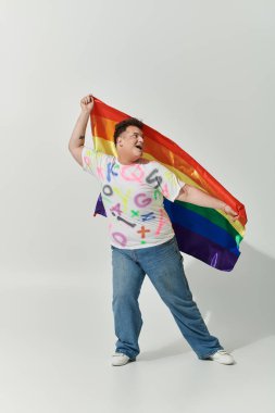 A queer person in colorful attire holds a rainbow flag, smiling brightly. clipart