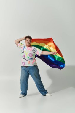 Renkli harflerle ve mavi kotla beyaz gömlekli tuhaf bir insan gökkuşağı bayrağıyla poz veriyor..