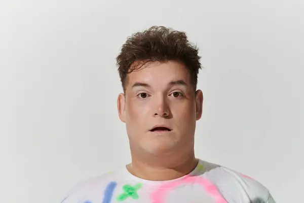 stock image A young queer person in a white shirt with splashes of color looks directly at the camera.