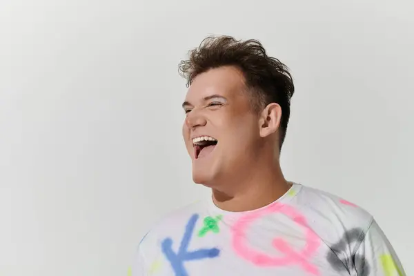 stock image A queer person wearing a white shirt with colorful spray paint designs laughs brightly at the camera.