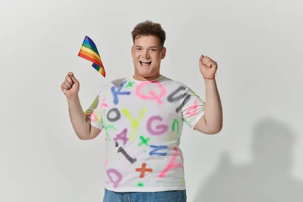 stock image A queer person with a colorful shirt proudly holds a rainbow flag and raises their fist in the air.