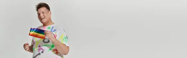 stock image A queer person in a colorful shirt holds a rainbow flag, smiling.