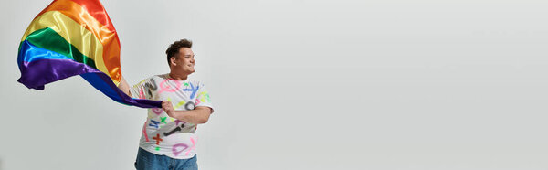 A queer person in colorful clothing holds a rainbow flag, smiling and looking to the side.