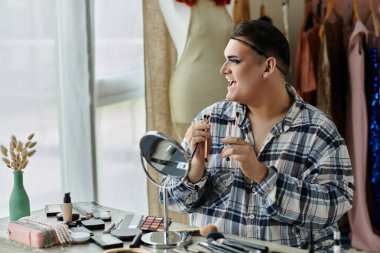 A man in a plaid shirt applies makeup in front of a mirror, surrounded by beauty products. clipart