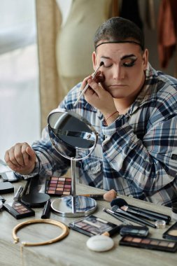 Eşcinsel bir insan, çeşitli makyaj aletleri ve ürünleriyle çevrili bir aynanın önünde göz makyajı yapar..