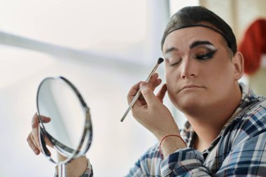 Eşcinsel bir insan göz farı sürer, yansıması el aynasında görünür..