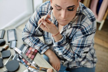 Ekose gömlekli tuhaf insan makyaj uygular. Etrafı aletlerle çevrili, aynaya bakar.