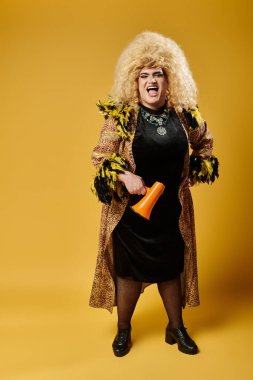 Parlak sarı saçlı ve kalın elbiseli bir travesti stüdyo ortamında bir megafon tutuyor..