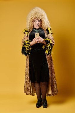 Şık giyimli bir travesti sarı bir arka planın önünde duruyor ve telefonlarına bakıyor..