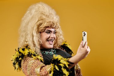 Parlak sarı peruklu bir travesti sarı arka planda bir selfie için poz verir..
