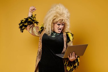 Büyüleyici giyinmiş bir travesti, elinde dizüstü bilgisayarla havaya bir yumruk indirir ve başarısını kutlar..