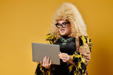 Şık tüylü ceketli bir travesti parlak sarı bir arkaplana karşı dizüstü bilgisayar kullanır..