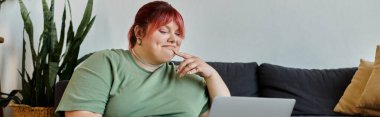 Pembe saçlı, iri yarı bir kadın evinde bir koltukta oturuyor ve dizüstü bilgisayara bakarken gülümsüyor..