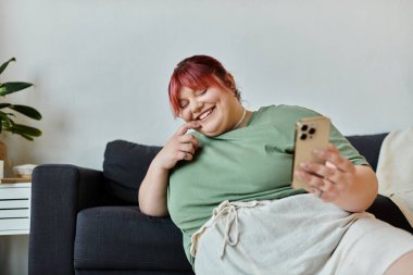 A woman laughs while sitting on a couch at home, using her phone. clipart