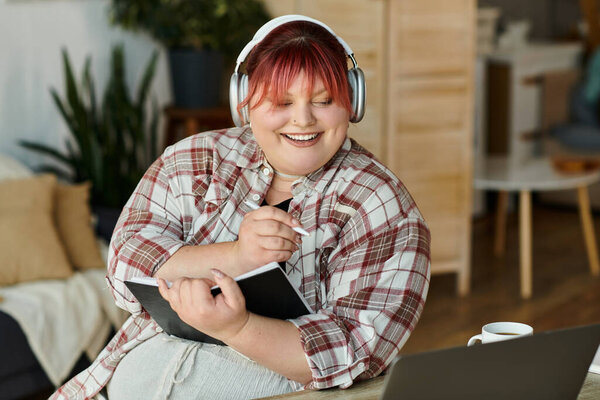 A woman in headphones writes in a journal, smiling at the camera.
