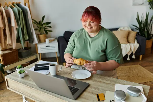 stock image A woman with pink hair smiles as she eats a croissant at her home office desk.