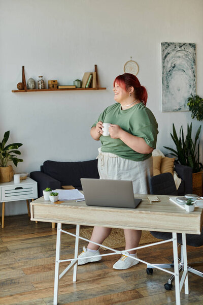 Женщина стоит у своего стола, держа кружку кофе и улыбаясь.