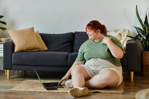 Женщина plus-size с рыжими волосами сидит на стуле, улыбается и пользуется ноутбуком.