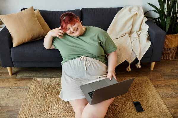 Женщина сидит на ковре и улыбается, когда пользуется ноутбуком..