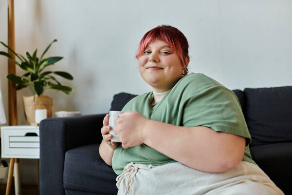 Женщина удобно сидит на диване, держа кружку, с спокойным выражением лица.