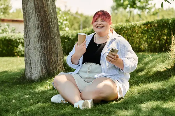 stock image A plus-size woman sits smiling under a tree, enjoying her coffee and looking at her phone.