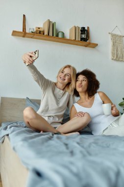 Çok kültürlü lezbiyen bir çift, rahat ev kıyafetleri içinde yataklarında selfie çekiyor..